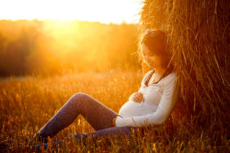 Young Pregnant Woman Sitting by the Haystack at Sunset and Embracing her Belly. 7 Month Pregnancy. Maternity Concept. Toned Photo.の写真素材