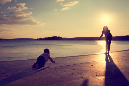 Silhouette of a Little Baby Crawling to His Mother on the Shore near Sea at Sunset. Active Child Playing and Family Love Concept. Toned Cross Processed Photo with Copy Space.の写真素材