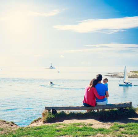 Family with Little Baby Sitting on a Bench near the Sea. Summer Vacation and Happy Parenting Concept. Toned Photo with Copy Space.の写真素材