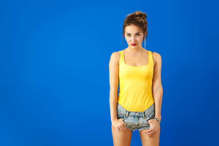 Sexy Summer Hipster Girl in Yellow Top and Jeans Shorts Posing at Blue Background. Trendy Street Style Fashion Outfit. Copy Space.の写真素材