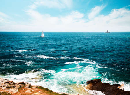 Summer Seascape with Transparent Clear Blue Water. Lighthouse and Sailing Yacht on Horizont. Sea Vacation Concept. Copy Space Background. Toned and Filtered Photo.の写真素材