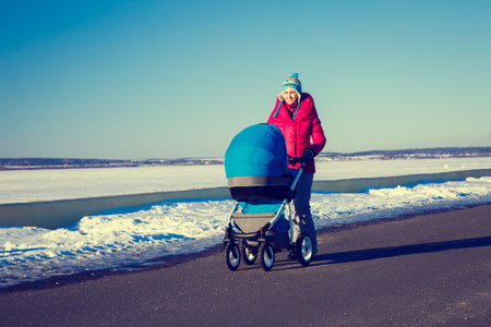 Happy Mother with Baby Stroller Walking in Winter Park and Enjoying Sunny Day. Snowy Background....