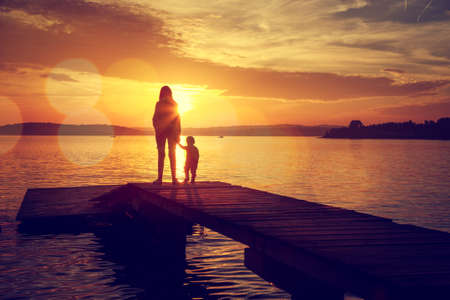 Silhouettes of Young Mother and Her Little Baby Standing on the Pier and Watching Sunset by the Lake. Happy Family Lifestyle. Motherhood and Childhood. Mom and Son. Toned Photo with Bokeh. Copy Space.の写真素材