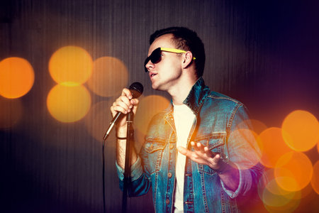 Young Guy in Sunglasses and Jeans Jacket Singing with Microphone. Singer Performance. Concert and Music Concept. Dark Background. Toned Photo with Bokeh.の写真素材