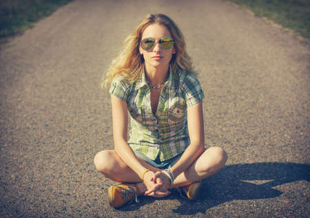 Street Style Hipster Girl Sitting on the Road. Fashion Woman in Sunglasses Outdoors in Summer. Beautiful Female Teenage. Toned Cross Processed Photo with Copyspace.の写真素材