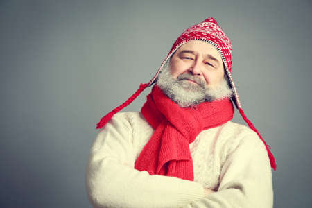 Portrait of Serious Old Man with Beard in Funny Red and White Winter Clothes on Gray Background. Modern Mature Man Concept. Toned Photo with Copy Space.の写真素材
