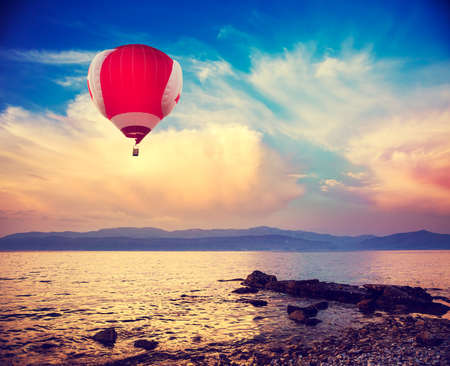 Hot Red Air Balloon Flying over Sea at Sunset. Blue Sky Background with Beautiful Clouds. Pebble Beach and Evening Seascape. Coastline and Mountains on the Horizon. Toned and Filtered Photo.の写真素材