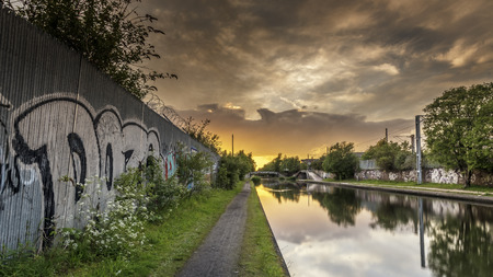 A graffiti covered wall, alongside a calm, flat canal, as the sun sets on a summers day.  the clouds and sky are reflecting in the still, flat waterの写真素材