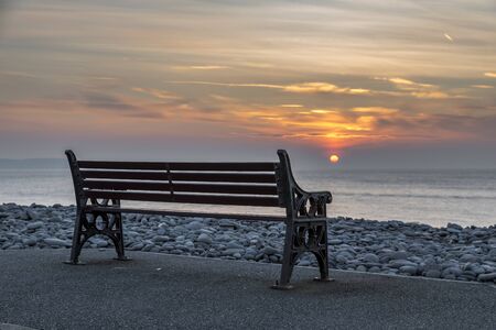 An empty bench, at the see shore, with a fiery, colourful sun setting into the oceanの写真素材