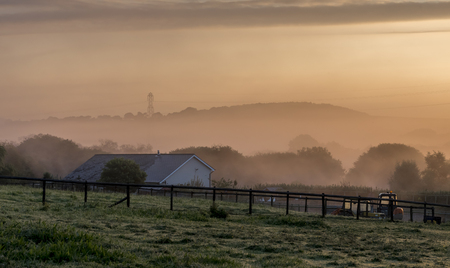 Autumn dawn, with an orange light through the early morning mist, looking over a rural farm scene to distance hillsの写真素材