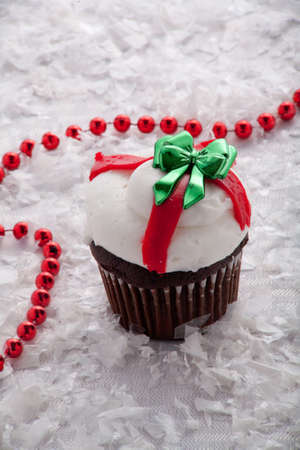 Christmas Chocolate Cup Cake With White Frosting and Green Bow Surrounded by Red Beads and Snowの写真素材