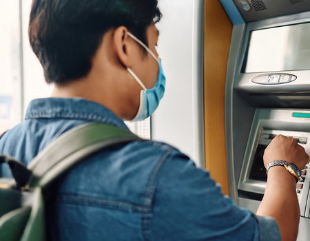 Young Asian man wearing face mask using automatic teller machine to withdraw money from credit card.の素材