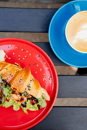 Croissant sandwich with cream cheese, salmon and arugula on a red plate on the terrace of a summer cafe, close-up. Healthy breakfast concept.の写真素材
