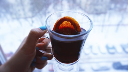 Woman holding mulled wine with spices and citrus fruit in her hand, close up in front of the window...