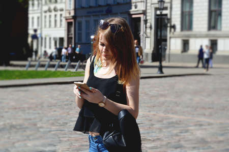 Beautiful young woman with blonde hair messaging on the smart-phone at the city street background. Pretty girl having smart phone conversation in sun flare.の写真素材