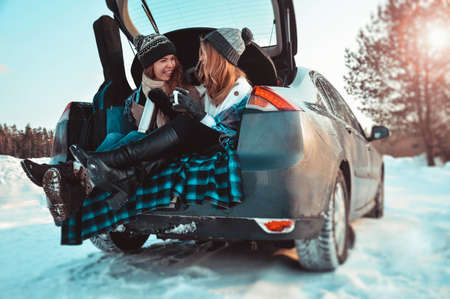 Happy friends in winter forest in the car. Two happy girls sit in the trunk of a car drinking coffee, talking and laughing.の写真素材