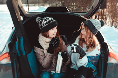 Happy friends in winter forest in the car. Two happy girls sit in the trunk of a car drinking coffee