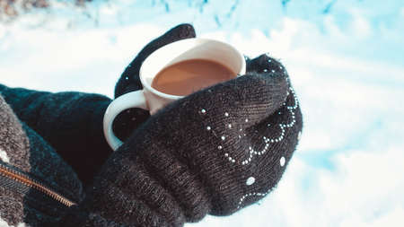 Warm cup of hot coffee warming in the hands in mittens. Womens hands on the background of snow, winterの写真素材