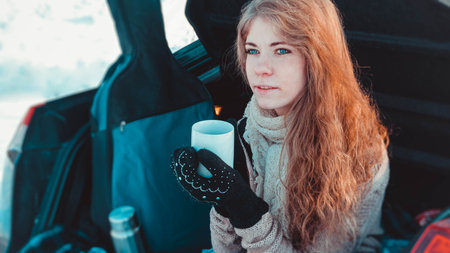 Happy girl in knitted clothes, scarf and plaid sitting on truck of car in winter forest with a cup...