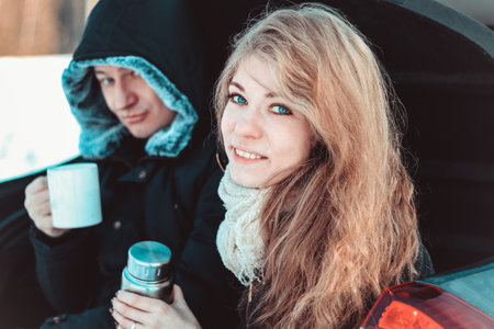 A man and a woman on truck of the car. A happy couple, wrapped in a warm knitted blanket, drinking...