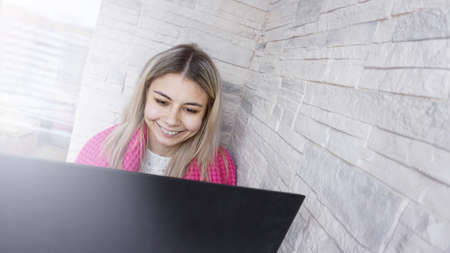 Beautiful smiling young woman with a laptop and warm plaid on a white brick backgroundの写真素材