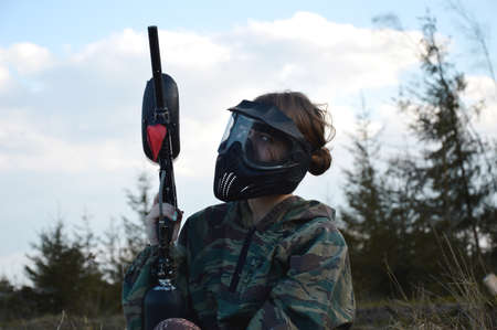 Paintball sport player girl in protective camouflage uniform and mask with marker gun outdoorsの写真素材