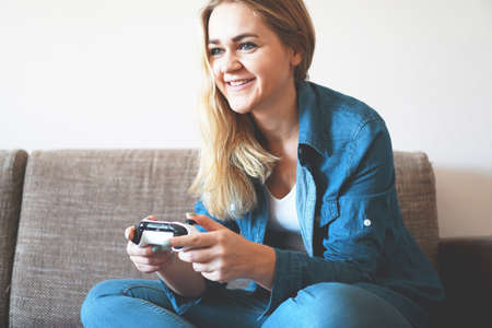 Girl gamer plays with a wireless gamepad while looking at the screen in front of her. Young blonde girl smiles and enjoys playing video games console.の写真素材