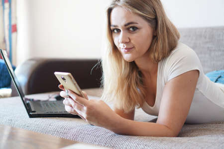 Attractive young woman relaxing on a sofa at home and using a touch screen smart phone to socialize and network from homeの写真素材