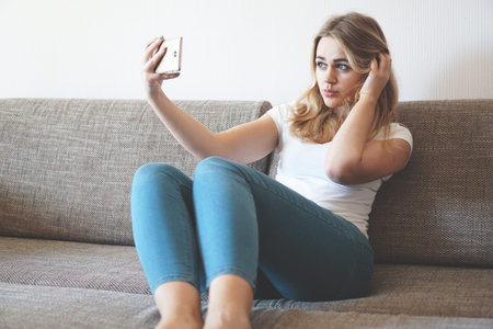 Shot of a happy young woman taking selfie with her cellphone while sitting at living roomの写真素材