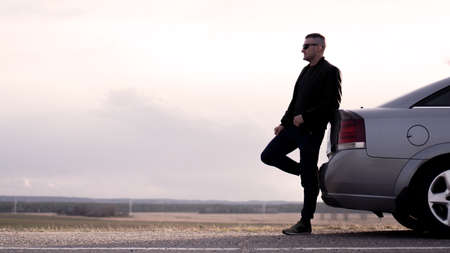 Sde view of young businessman in eyeglasses sitting on car trunk at nature backgroundの写真素材
