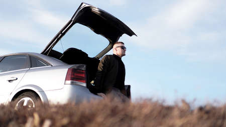 Sde view of young businessman in eyeglasses sitting on car trunk at nature backgroundの写真素材