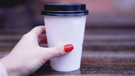 Cup of coffee on a wooden table in female hands. Paper cup Plastic cap. Beautiful hands with red nailsの写真素材