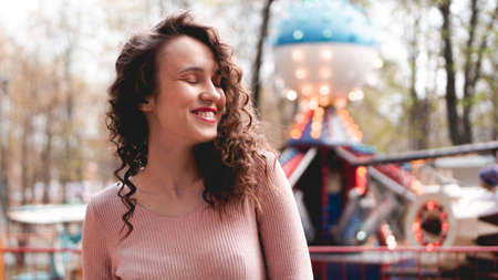 Girl chilling in amusement park in weekend morning. Laughing good-humoured female model with curly hair standing near carousel.の写真素材