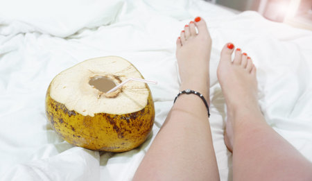 Female bare feet on a white sheet, top view, closeup. Coconut cocktail near the feet. Hotel by the...