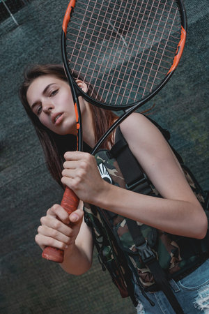Adorable woman holds a tennis racket, fashionable model, with long, brunette hair in camouflage vest...