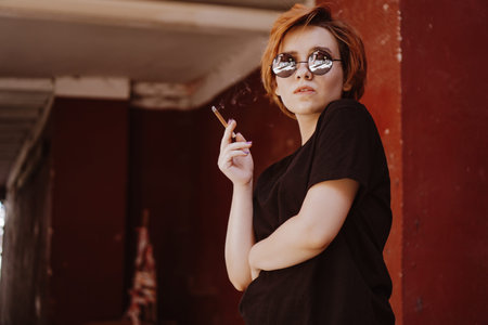 Millennial cool pretty girl with short red hair and mirror sunglasses smoking cigarette in the old...