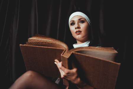 Beautiful young nun in religion black suit holds Bible and posing on camera with big book on a black background. Close-up. Religion concept.の写真素材
