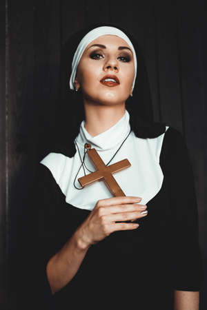 Sexy nun prays indoor . Beautiful young holy sister. Young beautiful nun with a cross in a robe in a black interior.の写真素材