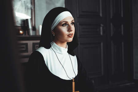 Sexy nun prays indoor . Beautiful young holy sister. Young beautiful nun with a cross in a robe in a black interior.の写真素材