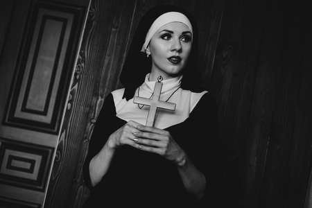 Sexy nun prays indoor . Beautiful young holy sister. Young beautiful nun with a cross in a robe in a black interior. Black and whiteの写真素材