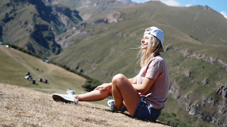 The girl in a cap and shorts in the mountains sits on the grass. Hike in the mountains, resting on a...
