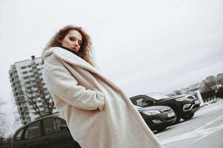 A girl with red curly hair in a white coat poses on outdoor parking in cold autumn. City Style - Urbanの写真素材