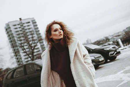 A girl with red curly hair in a white coat poses on outdoor parking in cold autumn. City Style - Urbanの写真素材