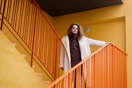 A girl with red curly hair in a white coat poses on the yellow orange parking stairs. City Style -...