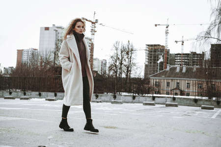 A girl with red curly hair in a white coat poses on outdoor parking in cold autumn. City Style - Urban. City construction in the backgroundの写真素材