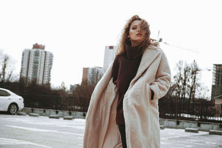 A girl with red curly hair in a white coat poses on outdoor parking in cold autumn. City Style - Urbanの写真素材