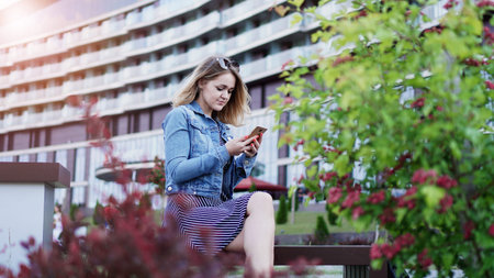 Girl texting on the smart phone sitting in a park - city building on the background