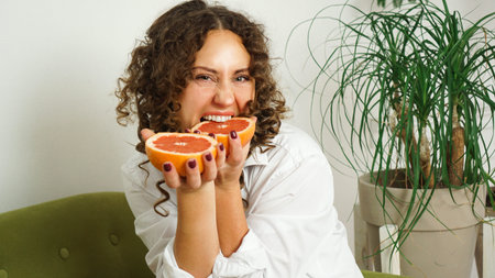Beautiful woman bites grapefruit. Middle-aged woman with curly hair at home - light room. The...