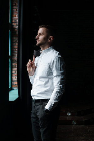 View in profile of a young businessman, dressed in a white shirt standing near the window on a dark...