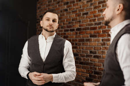 Stylish confident young man looking at himself in mirror. Fitting a suit in a store or sewing studio - loft interiorの写真素材